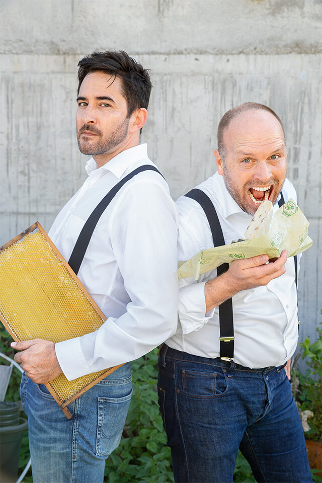 Zwei Männer, einer hält ein Brett Honigwaben, der andere möchte in einen Käse beißen, der in eine Bienenwachspapierverpackung eingewickelt ist.