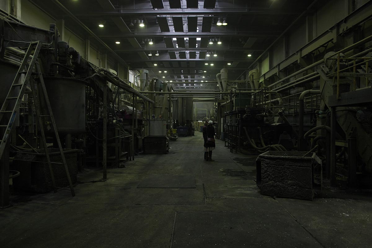 In einer dunklen Halle stehen zwei Personen umgeben von einem Rohr- und Förderleitungssystem.