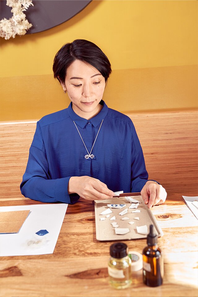 Eine Asiatin sitzt an einem Tisch und legt sich die Scherben eines zerbrochenen Porzellangeschirrs zurecht. Auf dem Arbeitstisch stehen diverse Fläschchen.