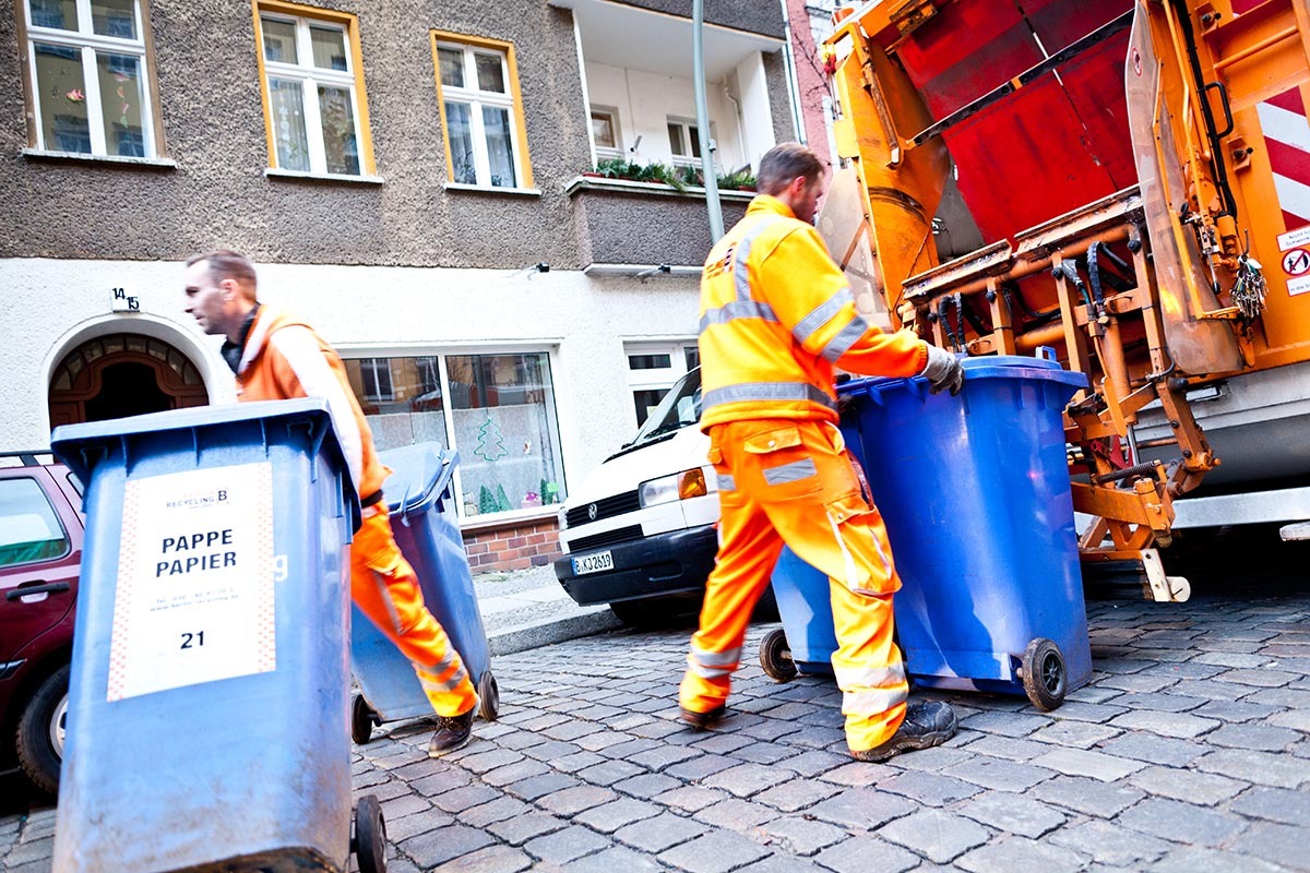 Zwei Männer in orangener Arbeitskleidung bringen blaue Papiertonnen zum Entleeren zum Müllfahrzeug und wieder zurück.
