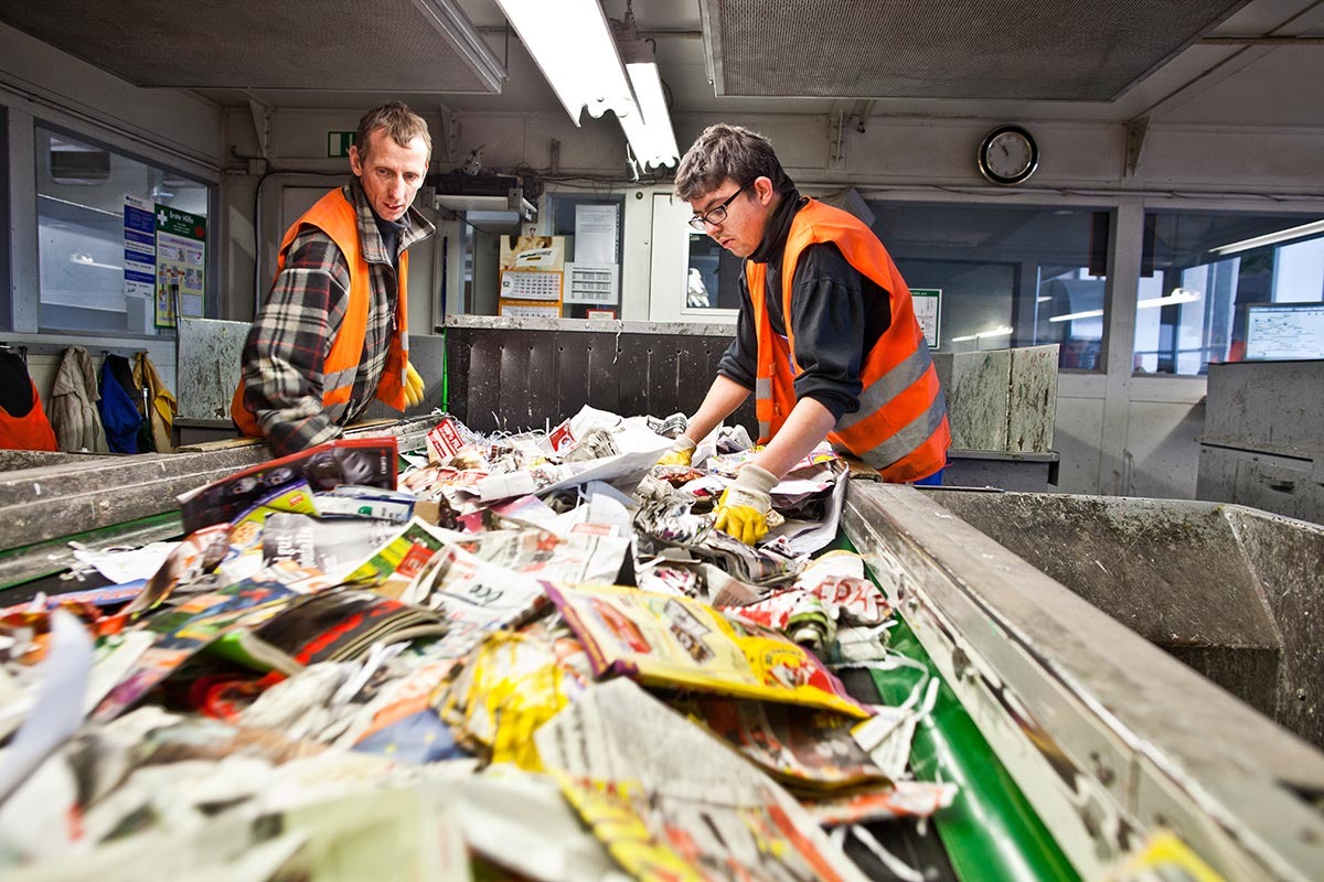 Zwei Männer in orangener Arbeitskleidung sortieren Altpapier am Förderband.