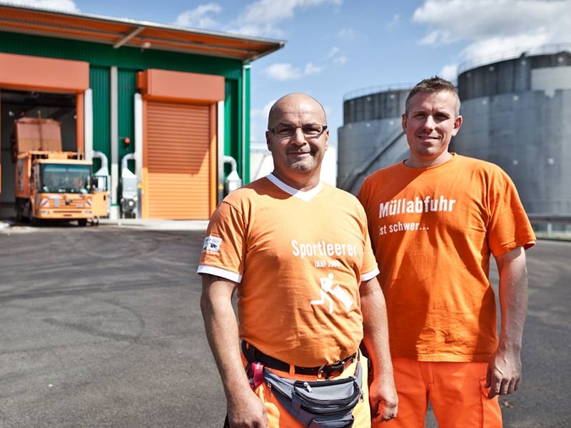 Zwei Männer in oranger Arbeitskleidung posieren vor einer Müllabladehalle. Im Hintergrund ein Müllfahrzeug beim Müllabladen.