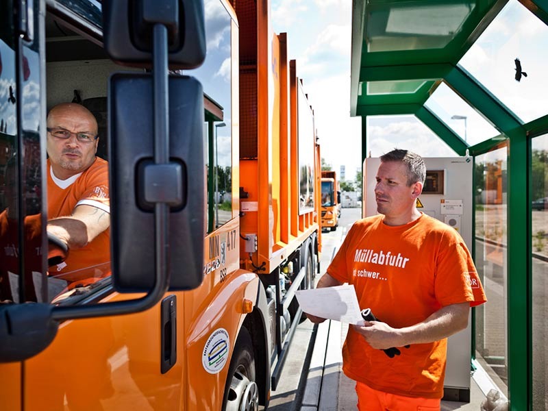 Zwei Männer in oranger Arbeitskleidung wiegen das Müllfahrzeug: Einer sitzt im Auto, der andere steht mit Papieren in der Hand vor dem Auto.