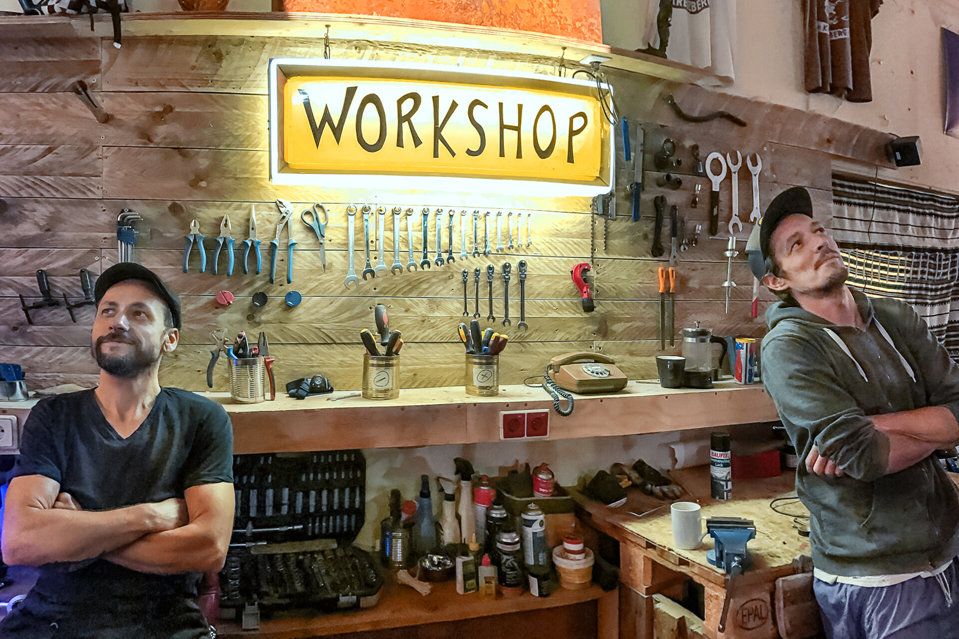 Zwei Männer stehen lächelnd und mit verschränkten Armen in einer Fahrradwerkstatt. Im Hintergrund Werkzeuge und eine Leuchtbox, auf der „Workshop“ steht.